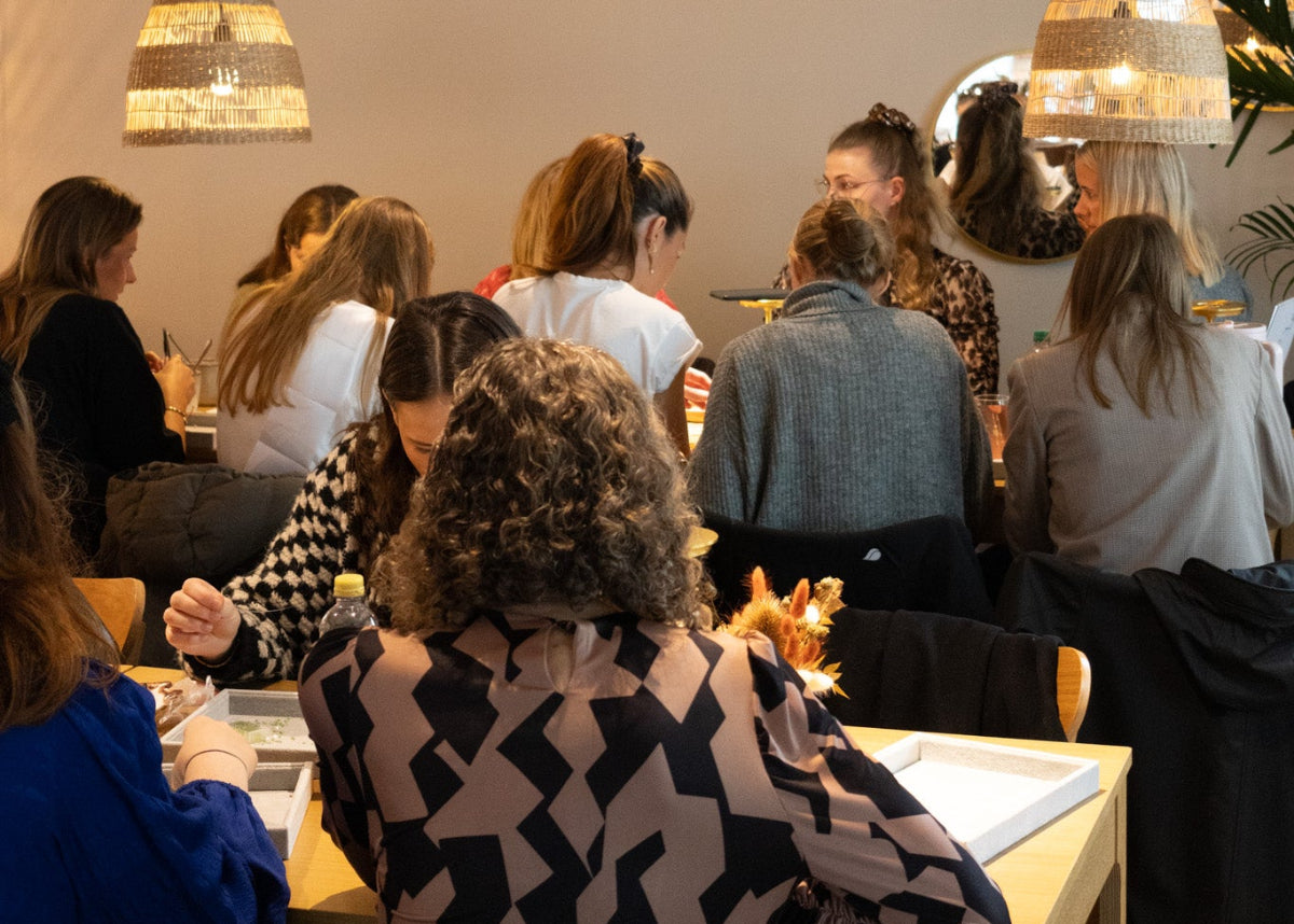 En gruppe kvinder sidder samlet ved borde i Smyk&Hyg i Vejle og laver smykker sammen i en varm og hyggelig atmosfære med dæmpet belysning.