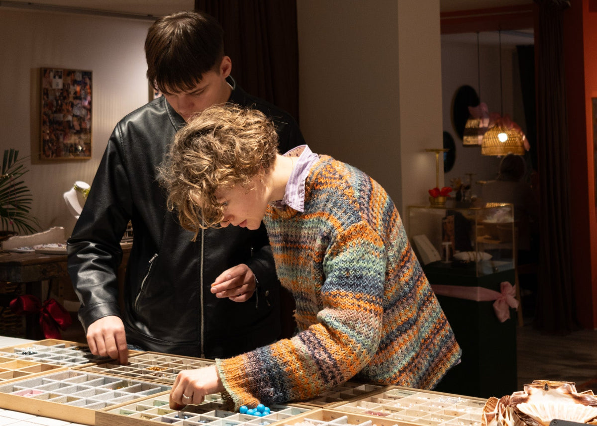 To unge personer står ved et bord i Smyk&Hyg i Vejle og udvælger perler fra sorteringsbakker i en hyggelig og kreativ atmosfære med varmt lys.