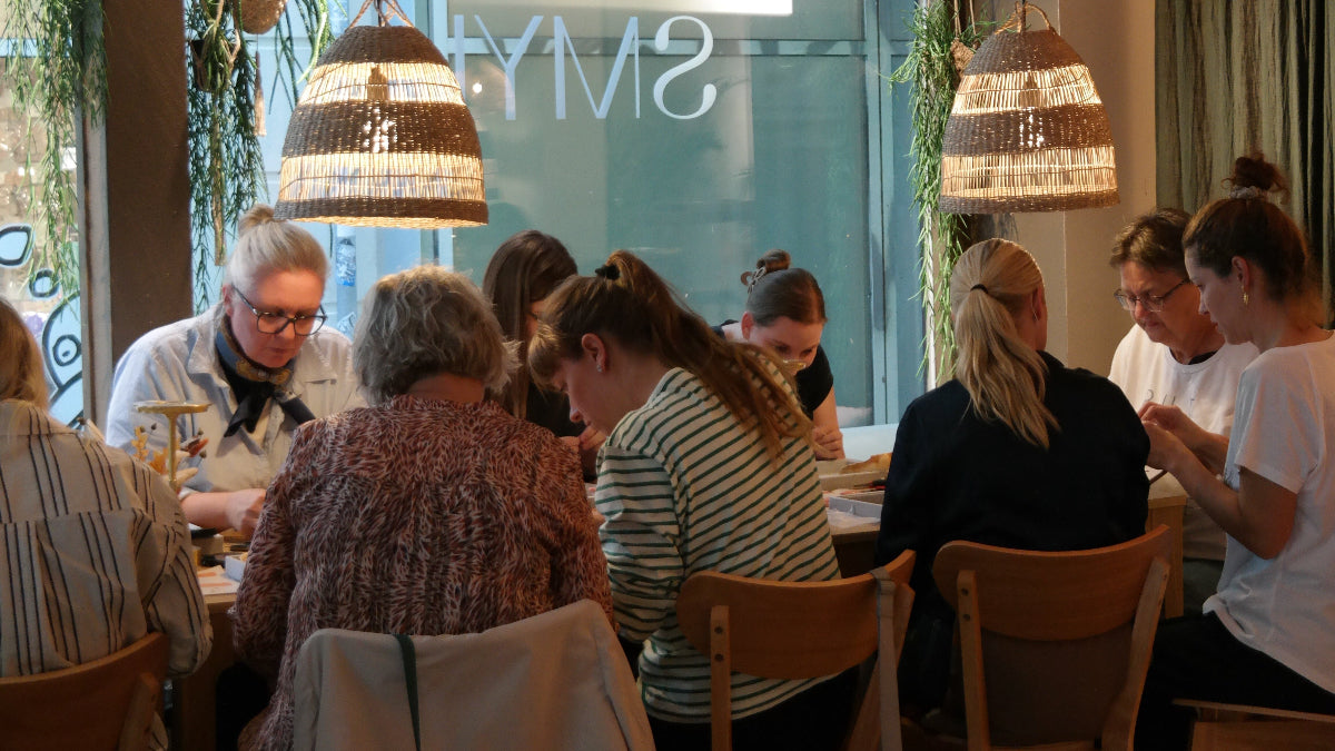 Gæster sidder samlet omkring et bord hos Smyk&Hyg i Vejle og laver deres egne smykker i en hyggelig og kreativ atmosfære.