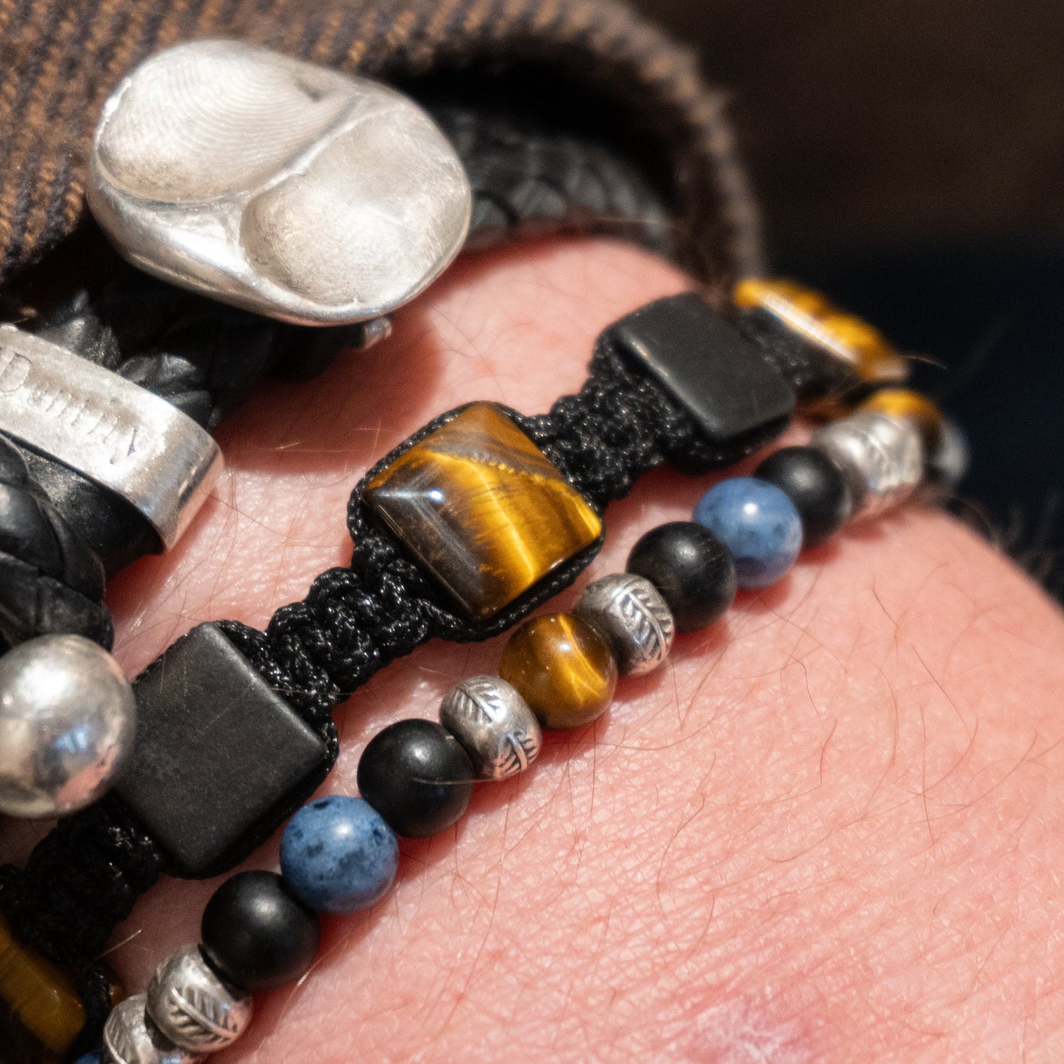 Læderarmbånd på en herrearm med håndlavet sølvvedhæng præget med fingeraftryk samt et knyttet armbånd i sort snor med sorte og brune perler. 