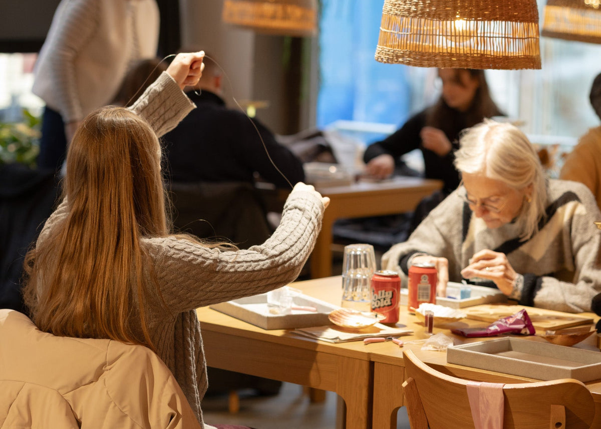 Kvinder i forskellige aldre sidder ved et bord i Smyk&Hyg i Vejle og laver smykker sammen i en hyggelig og kreativ atmosfære med varme lamper og perler på bordet.