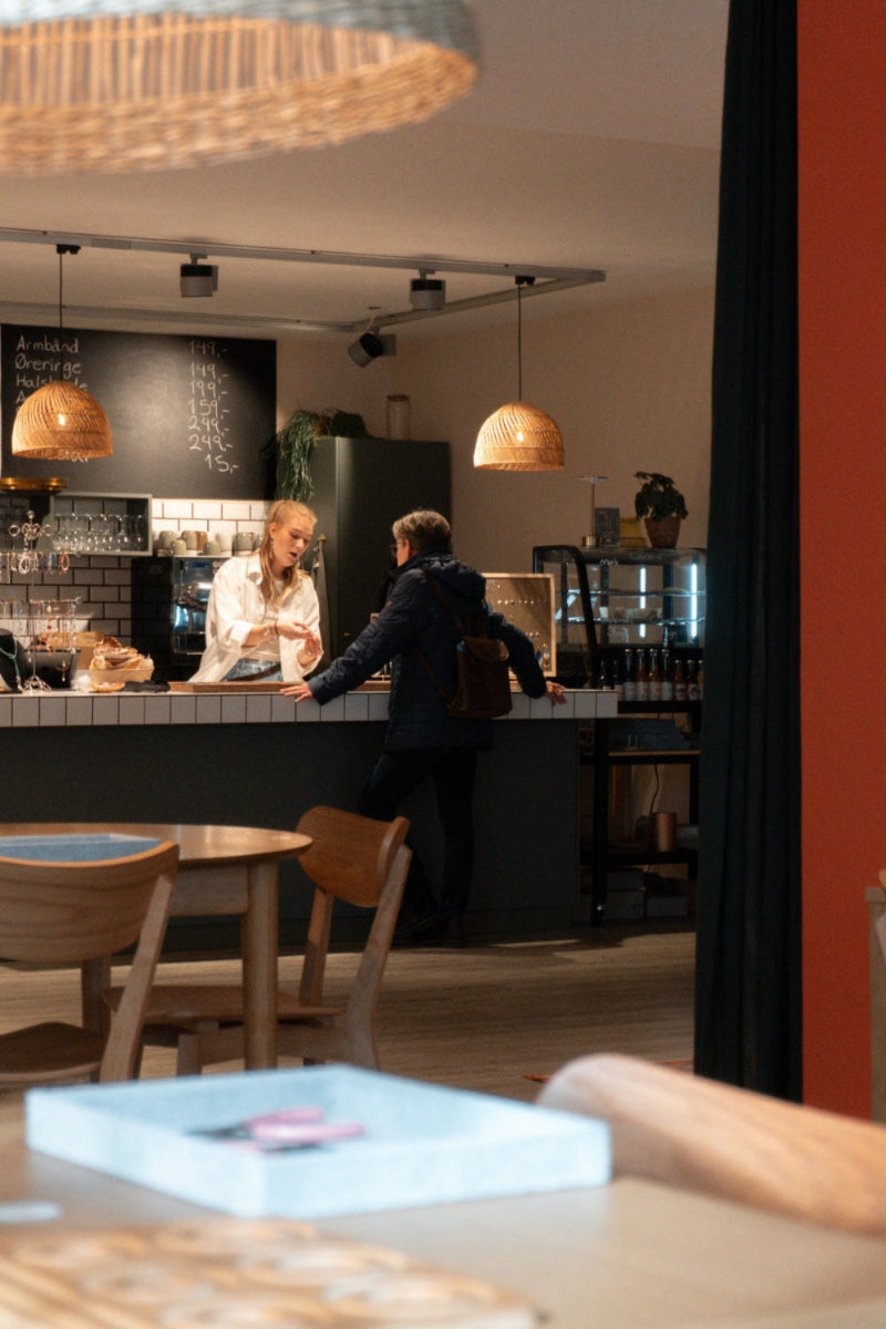 Kunde og medarbejder i samtale ved kaffedisken i Smyk & Hygs hyggelige smykkecafé i Vejle med varme lamper og afslappet atmosfære