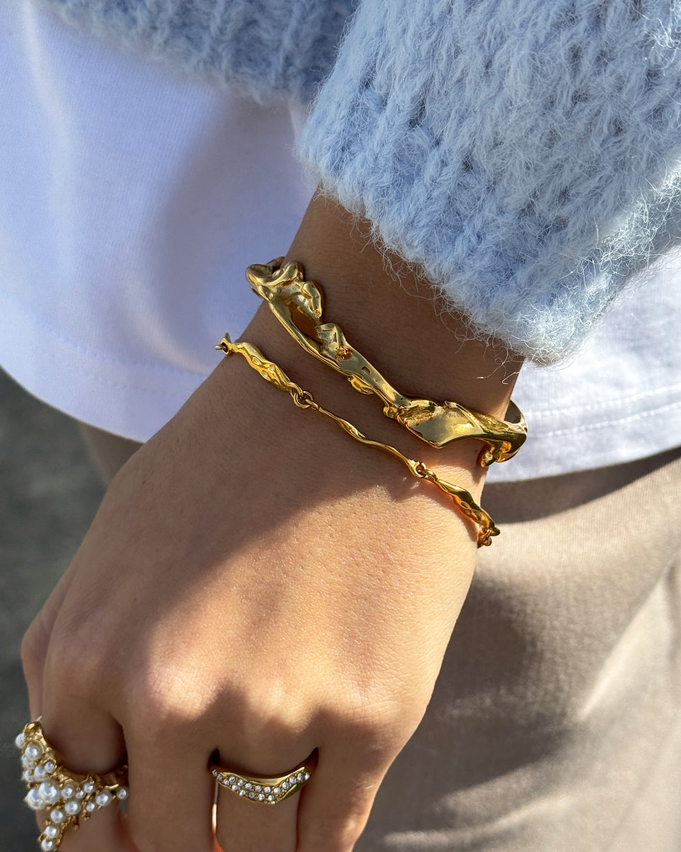 Hånd med guldfarvede armbånd og ringe fra Bay by Aqua Dulce – vandfaste smykker hos Smyk&Hyg i Vejle