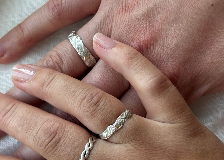 En mandehånd med en vielsesring i sølv med fingeraftryk. Oven på mandehånden hviler en kvindehånd, også med en vielsesringe i sølv med fingeraftryk